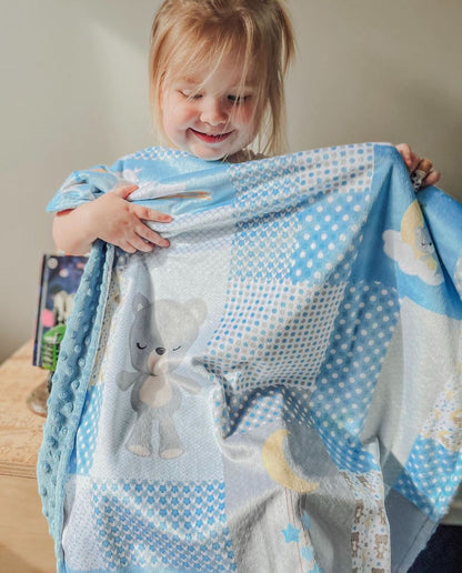 A smiling toddler holding up a Love Always minky blanket in a blue bear and moon patchwork print, showing how big it is